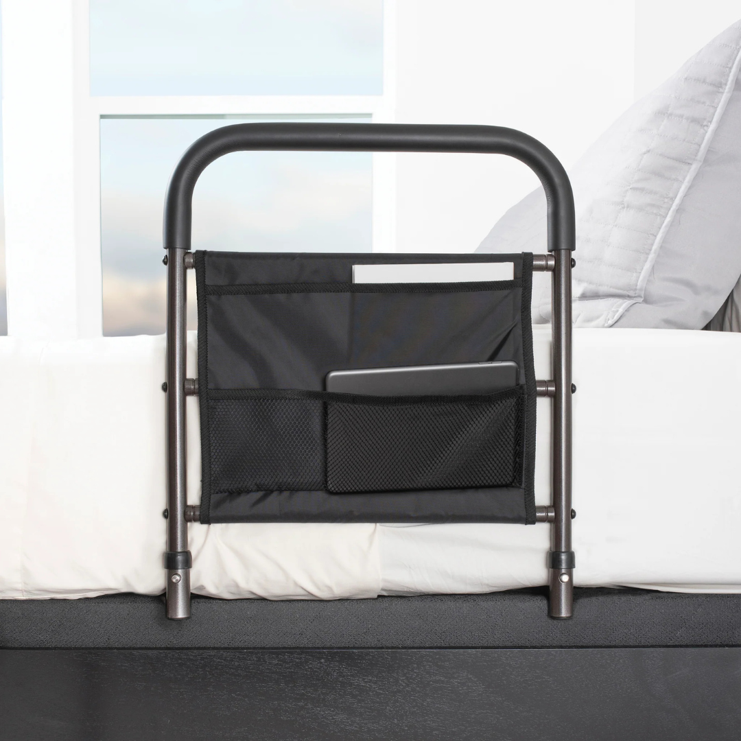 The Stander Wonder Bed Handle, a black bed rail with a fabric organizer pocket, attaches to your bed for added bedside safety and holds items like a tablet. The background features a white bed, gray pillow, and window.